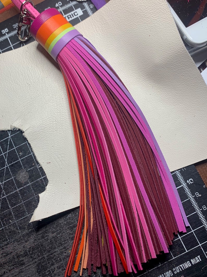 Op de afbeelding: Een veelkleurige leren kwast sleutelhanger met een regenboogstreepjespatroon. De kwast is gemaakt van dunne leren stroken in tinten roze, paars, oranje en rood.