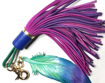 May include: A teal blue leather feather charm with a gold clasp attached to a purple and pink leather tassel with a blue leather strap.