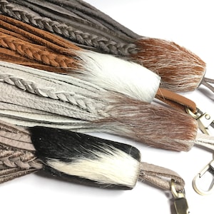 Puede incluir: Cuatro llaveros de cuero con correas de cuero trenzadas y pompones de piel en marrón, blanco y negro.