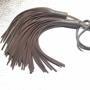 Puede incluir: Un borla de cuero marrón con una larga franja fina. La borla está unida a un cordón de cuero marrón.