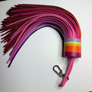 Op de afbeelding: Een kleurrijke leren sleutelhanger met een kwast gemaakt van meerdere tinten roze, paarse en rode leren stroken. De kwast is bevestigd aan een roze leren band met een zilveren sleutelring.