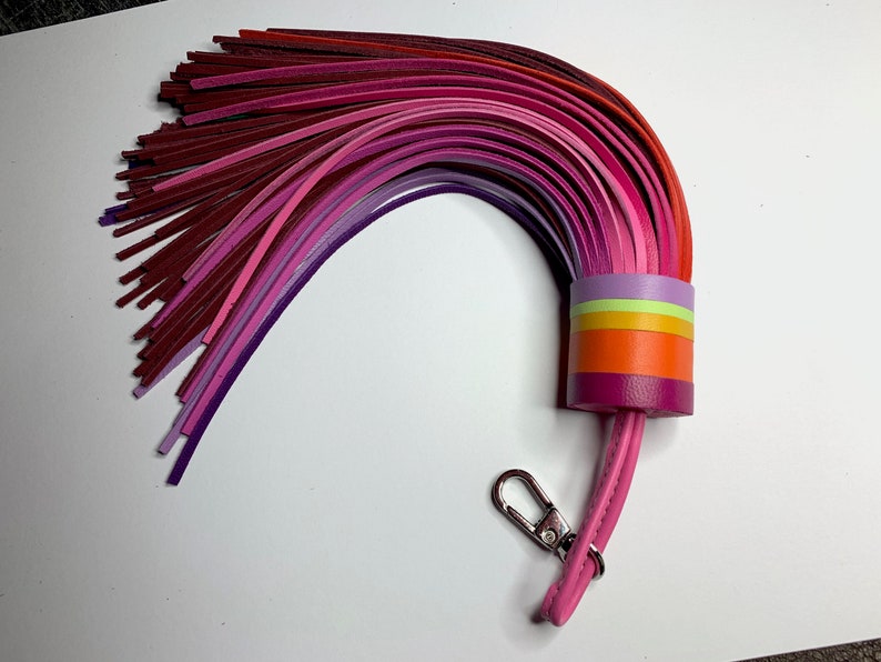 Op de afbeelding: Een kleurrijke leren sleutelhanger met een kwast gemaakt van meerdere tinten roze, paarse en rode leren stroken. De kwast is bevestigd aan een roze leren band met een zilveren sleutelring.