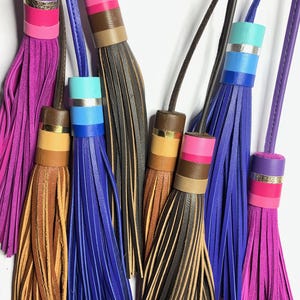 Puede incluir: Colección de borlas de cuero coloridas en tonos morado, rosa, azul, marrón y negro. Cada borla presenta una parte superior cilíndrica con bandas de colores contrastantes y un fino cordón de cuero para colgar. Fondo blanco.