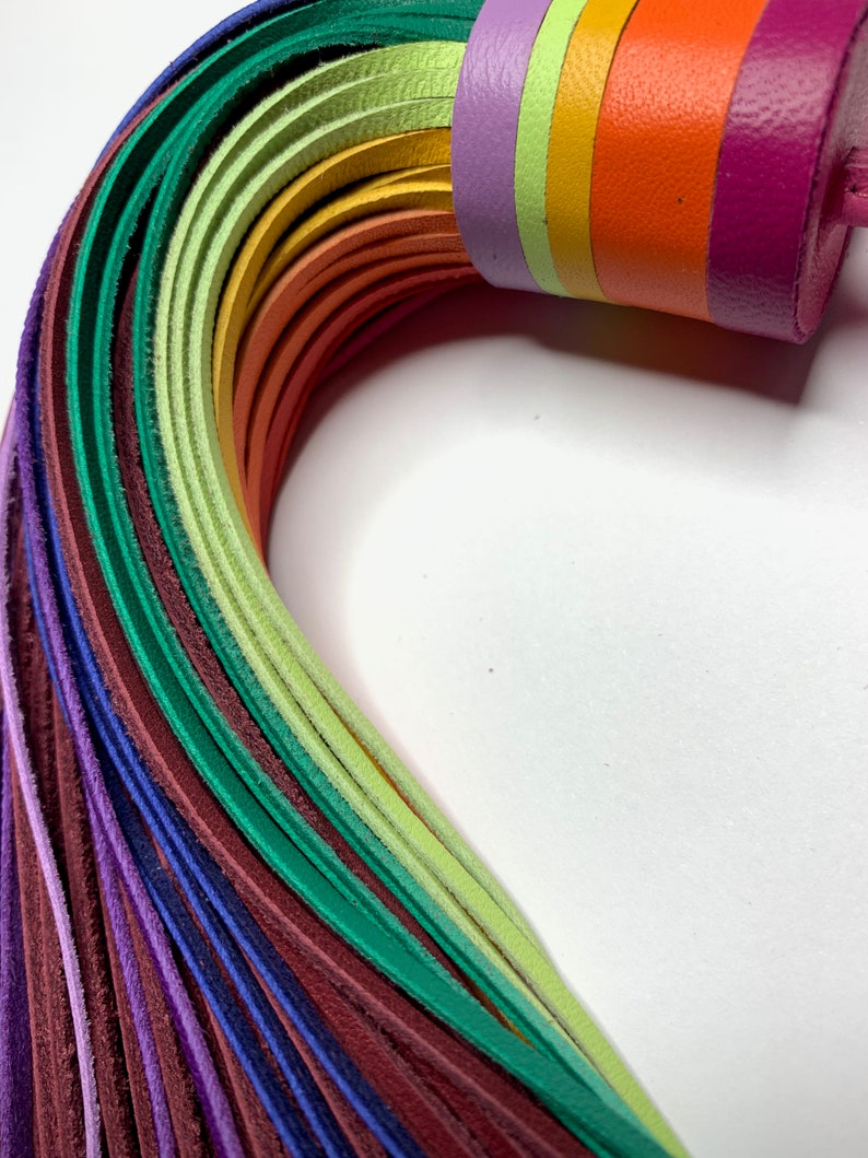 Op de afbeelding: Een regenboogkleurige leren zweep met een veelkleurige handgreep. De zweep is gemaakt van veel dunne leren stroken in tinten paars, groen, geel, oranje en roze.