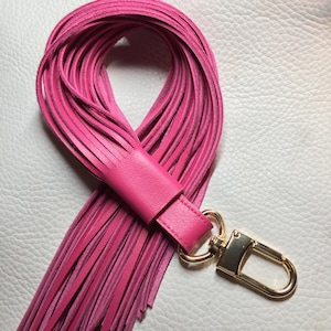 Puede incluir: Un llavero de cuero rosa con un largo fleco de borlas y un anillo de llave de metal dorado.