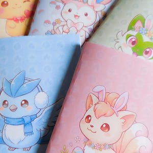 May include: A collection of notebooks featuring colorful illustrations of cartoon characters. The notebooks have different colored covers, including yellow, blue, pink, and green. The illustrations include cute animals with floral and Easter-themed decorations.