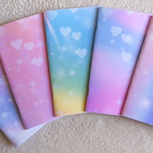 Può includere: Cinque quaderni con copertine sfumate pastello. Ogni quaderno presenta una diversa combinazione di colori, tra cui rosa, blu, viola e giallo, con motivi a cuori bianchi e glitter. I quaderni sono aperti, rivelando pagine bianche.