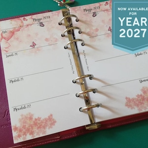 Könnte beinhalten: Ein rosa-roter Planer mit einem Metallordner, geöffnet auf einer Seite mit Blumen- und Schmetterlingsmotiven. Die Seite ist mit "Maggie 2019" und "Maggie 2013" datiert. Der Text "NOW AVAILABLE FOR YEAR 2027" wird auf einem blau-weißen Banner angezeigt.