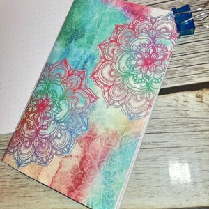 Può includere: Un taccuino con un design ad acquerello con due motivi mandala nei toni del rosso, rosa, blu e verde. Il taccuino è tenuto insieme da una clip blu. Il testo "2 - MANDALA" è visibile in basso.