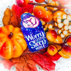 Puede incluir: Marcapáginas morado con un diseño de unicornio y el texto "Worry Less Sleep More". El marcapáginas está rodeado de follaje de otoño y pequeñas bayas naranjas y blancas.