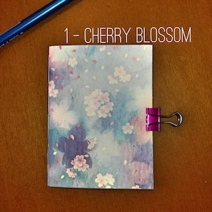 Può includere: Un piccolo taccuino con un motivo floreale di fiori di ciliegio, tenuto insieme da una clip rosa. Il taccuino ha uno sfondo in stile acquerello nei toni del blu, viola e rosa. Il testo "1 - CHERRY BLOSSOM" è visibile. Una penna blu e una rossa sono sullo sfondo.