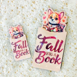 Könnte beinhalten: Zwei Herbst-Lesezeichen mit einem Cartoon-Corgi-Hund, der ein Buch hält. Das größere Lesezeichen hat einen glitzernden rosa und roten Hintergrund mit dem Text "Fall into a Book". Das kleinere Lesezeichen hat einen einfarbigen braunen Hintergrund mit dem gleichen Text.