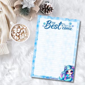 May include: Blue and white lined notepad with a snowflake pattern and the text "The Best is Yet to Come". A mermaid illustration is in the bottom right corner. The notepad is on a white fluffy surface.