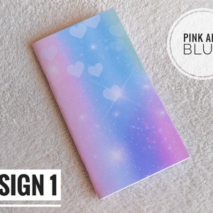 Può includere: Un quaderno con un design galassia pastello nei toni del rosa, blu e viola. Cuori e stelle bianche sono sparsi sulla copertina. Il testo "DESIGN 1" è in un riquadro bianco in basso a sinistra. Il testo "PINK AND BLUE" è in un cerchio bianco in alto a destra.