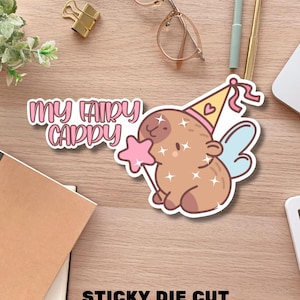 May include: A die-cut sticker featuring a brown capybara with light blue wings, wearing a yellow party hat with a pink heart, and holding a pink star wand. The sticker has a holographic glitter finish and the text "MY FAIRY CAPPY". It measures 8 x 5 cm.