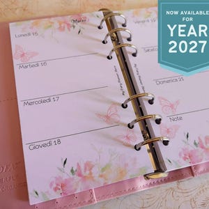 Puede incluir: Un organizador personal Filofax Butterfly rosa abierto en una página de planificador semanal. La página presenta diseños florales y de mariposas, con el texto "NOW AVAILABLE FOR YEAR 2027". El planificador está abierto a una semana de marzo.