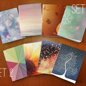 Può includere: Due serie di piccoli quaderni sono esposte su una superficie di legno. Il set 1 presenta copertine con un motivo floreale, una scena del tramonto, foglie autunnali e un disegno di fiocco di neve. Il set 2 include copertine con forme geometriche, un girasole e una scena notturna stellata.