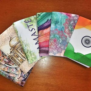 Può includere: Una collezione di quaderni colorati con vari disegni. Un quaderno presenta il testo "NAMASTE", mentre altri mostrano motivi di piume, mandala e una bandiera indiana. I quaderni sono disposti su una superficie di legno.