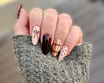 May include: A hand with long, oval-shaped nails. The nails feature various designs, including a bow, a tortoiseshell pattern, a plaid pattern, and polka dots. The color palette includes shades of brown, beige, and white. The nails are well-manicured and polished.