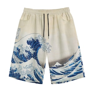 May include: Off-white shorts featuring a blue graphic print of a large ocean wave and a mountain. The shorts have an elastic waistband with black drawstring cords and two side pockets. The design includes Japanese text in a vertical rectangular box.
