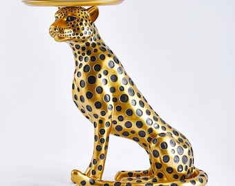 May include: A gold cheetah sculpture with black spots, holding a round gold tray on its back. The sculpture is a decorative accent piece for a home.