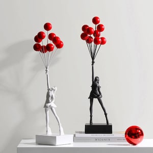 Peut inclure: Deux sculptures en noir et blanc d'une personne tenant des ballons rouges. Les sculptures sont sur une surface blanche avec un livre intitulé "KINFOLK" sur la surface.