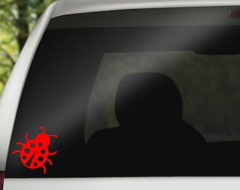 May include: A red ladybug decal is affixed to the rear window of a white vehicle. The ladybug design features a rounded body with black spots and antennae. The background shows a blurred view of trees.