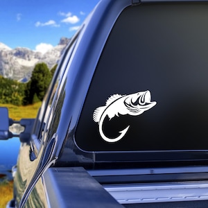 Könnte beinhalten: Weißes Vinyl-Abziehbild eines Großmaulbasses, der auf einen Angelhaken zuspringt. Der Aufkleber befindet sich auf der Heckscheibe eines schwarzen Trucks, mit Berg- und Seehintergrund.