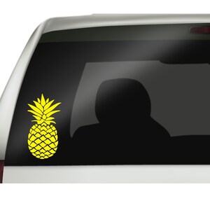 Op de afbeelding: Een gele ananassticker is bevestigd op de achterruit van een auto. Het ananasontwerp is effen van kleur, met een gedetailleerde textuur. De autoruit is getint en de achtergrond is een vage weergave van het interieur.
