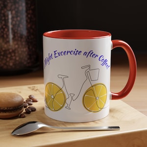 Puede incluir: Taza de cerámica blanca con interior y asa rojos. La taza presenta un diseño de bicicleta con ruedas amarillas y el texto "Night Excercise after Coffee" en azul. Una cuchara y un pastelito están en primer plano.
