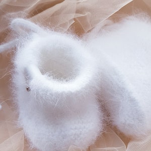 May include: A pair of white fuzzy baby booties on a beige tulle fabric background.