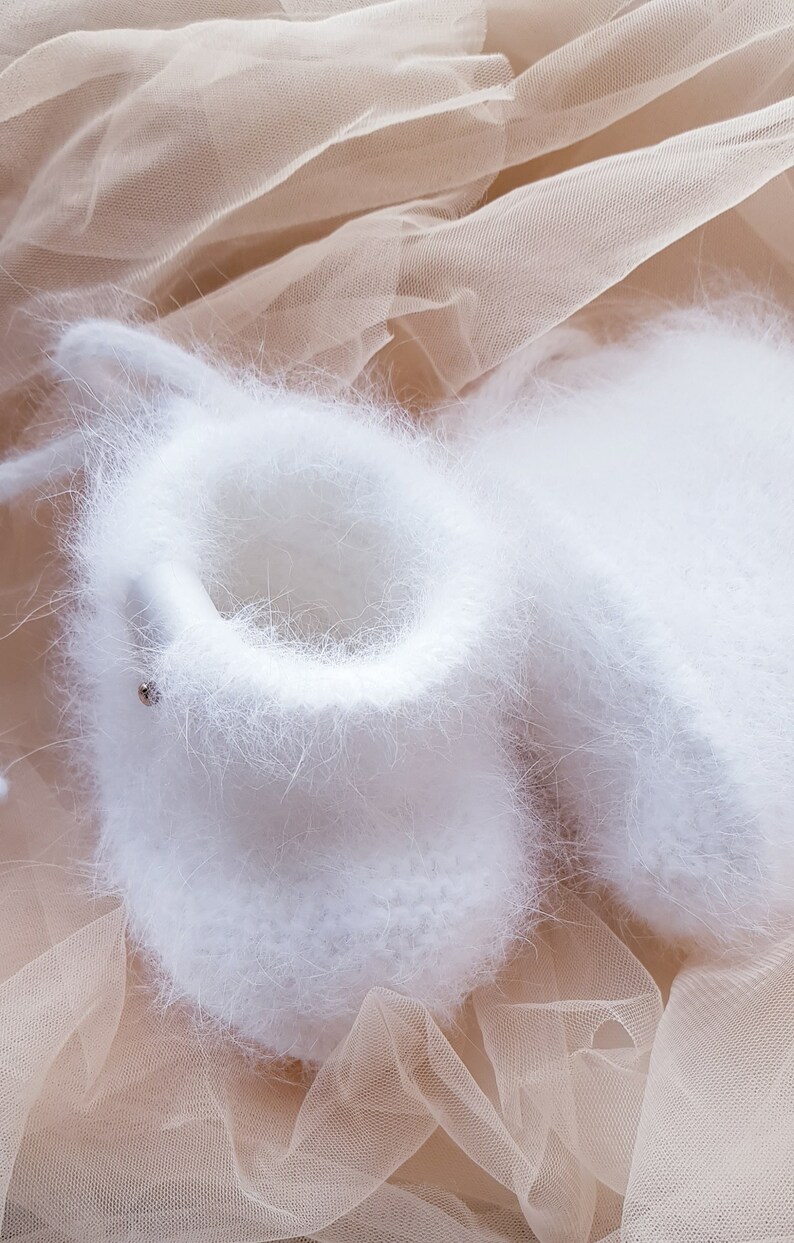 May include: A pair of white fuzzy baby booties on a beige tulle fabric background.