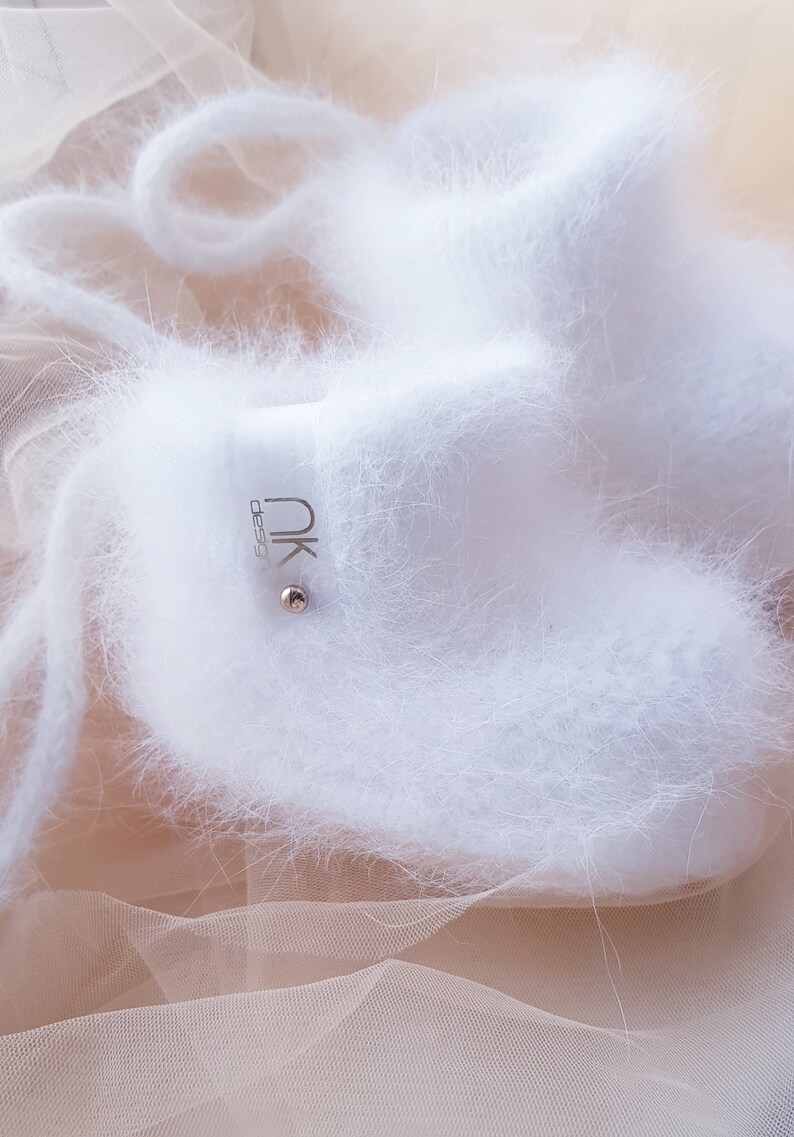 May include: White fuzzy baby booties with a silver tag that says "nk design". The booties are on a sheer peach fabric.