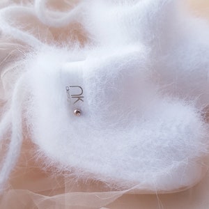 May include: White fuzzy baby booties with a silver tag that says "nk design". The booties are on a sheer peach fabric.