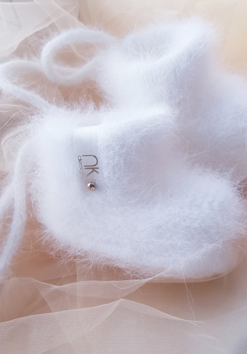 May include: White fuzzy baby booties with a silver tag that says "nk design". The booties are on a sheer peach fabric.