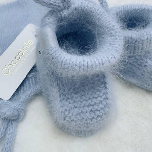 May include: A pair of light blue baby booties with a fuzzy texture. The booties are tied together with a matching ribbon. The booties have a tag that says "Chocolate Design & Studio".