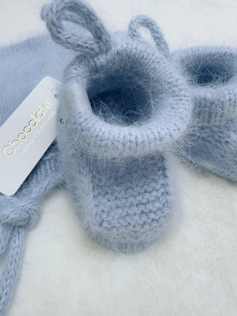 May include: A pair of light blue baby booties with a fuzzy texture. The booties are tied together with a matching ribbon. The booties have a tag that says "Chocolate Design & Studio".