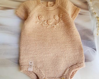 Könnte beinhalten: Ein hellpfirsichfarbener gestrickter Baby-Strampler mit kurzen Ärmeln und Holzknöpfen. Der Strampler hat ein Blumendesign auf der Brust und ein kleines Etikett an der Seite. Das Kleidungsstück ist aus weichem Garn gefertigt.