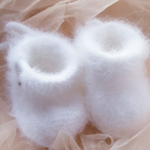 May include: A pair of white fuzzy baby booties on a beige tulle fabric background.