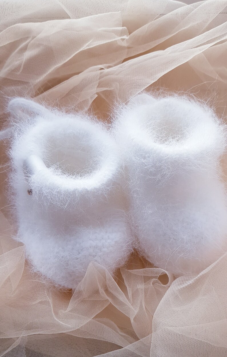 May include: A pair of white fuzzy baby booties on a beige tulle fabric background.