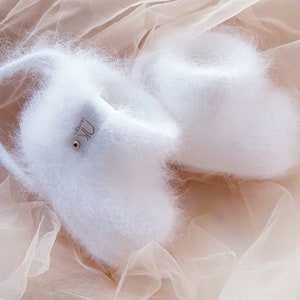 May include: Two white fluffy slippers with a gold logo that says "Uk Design" on one slipper. The slippers are on a light brown tulle fabric.