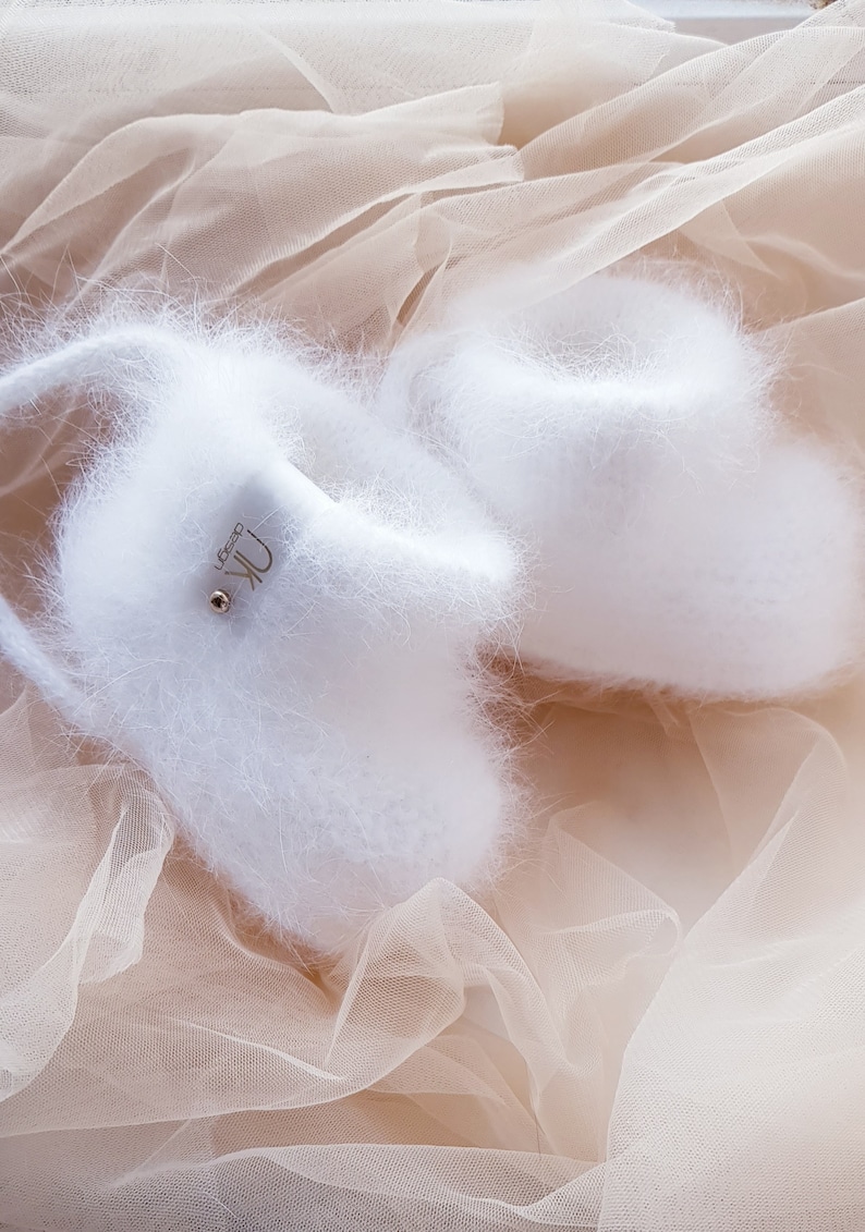 May include: Two white fluffy slippers with a gold logo that says "Uk Design" on one slipper. The slippers are on a light brown tulle fabric.