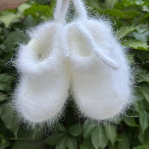 May include: White knitted baby booties with a fuzzy texture and a white string loop.