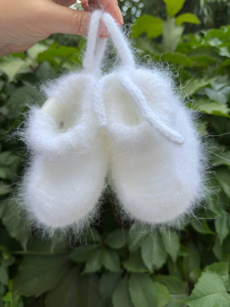 May include: White knitted baby booties with a fuzzy texture and a white string loop.