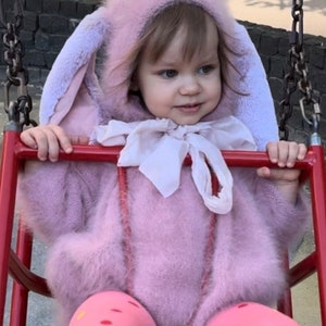 May include: A little girl wearing a pink fuzzy bunny costume with large ears, sitting on a red swing set.