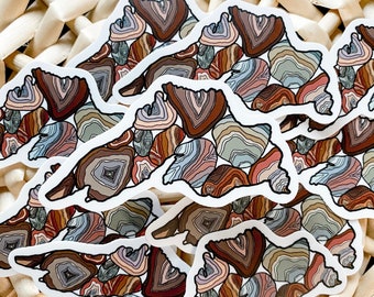 May include: A collection of stickers featuring a mountain range design filled with colorful, layered agate patterns. The stickers have a white border and showcase shades of brown, gray, blue, and red. The stickers are arranged on a woven, light-colored surface.