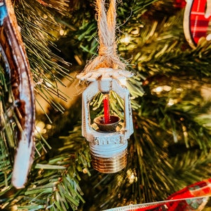 May include: A white and gold Christmas tree ornament shaped like a fire sprinkler head, suspended by a natural twine string. The ornament has a red element inside and is set against a backdrop of green pine needles and festive decorations.