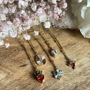 Könnte beinhalten: Eine Sammlung goldfarbener Halsketten mit verschiedenen Anhängern, darunter eine Erdbeere, ein Sonnenstrahl, ein quadratischer Edelstein, ein Schneeflocke und eine kleine Blume. Die Halsketten werden auf einer Holzoberfläche mit rosa und weißen Blumen im Hintergrund präsentiert.