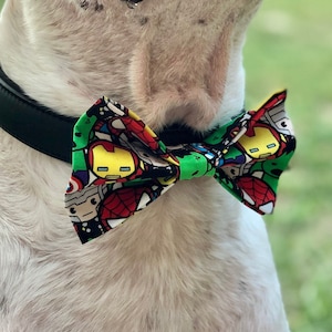 May include: A white and black spotted dog wearing a black collar and a colorful bow tie with a superhero theme. The bow tie features a variety of characters, including Iron Man, Captain America, and Thor.
