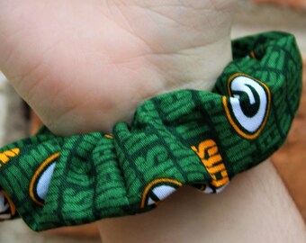 Puede incluir: Scrunchie de tela verde y amarilla con el logotipo y el texto de los Green Bay Packers. El scrunchie está en la muñeca de una persona.